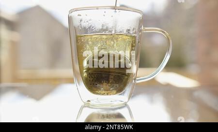 Gießen Sie heißes Wasser mit einer Teebälle in einen Glasbecher. Stockfoto