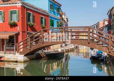 Braune hölzerne Fußgängerbrücke über den Kanal mit festgemachten Booten, rot-blauen Stuckhäusern und Schaufensterfronten, Burano Island, Lagune Venezianischer Seen, Venedig, Italien. Stockfoto