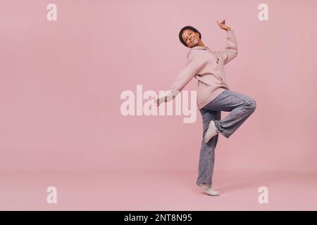 Junge, lächelnde schwarze Frau, die über pinkfarbenen Hintergrund tanzt und springt. Süßes glückliches Mädchen mit Jeans und Kapuzenpullover, das vor der Kamera posiert Stockfoto