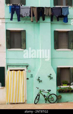 Grüne Stuckfassade mit weiß-gelb gestreiften Vorhängen über der Eingangstür und gewaschene Kleidung an Wäscheleine, Fahrrad, Burano, Italien. Stockfoto