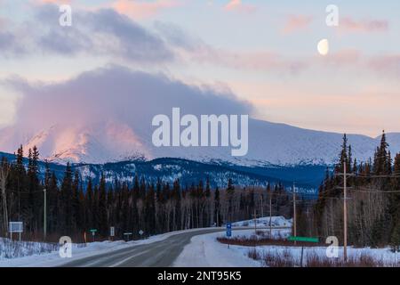 Pastellrosa Sonnenaufgang entlang des Alaska Highway in der Wintersaison mit Monduntergang über der Wildnis. Stockfoto