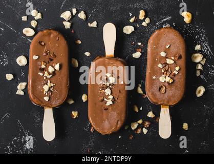 Milchschokolade Eis am Stiel mit Haselnüssen. Nahaufnahme. Eis am Stiel mit Schokolade, Stäbchen und schwarzem Steinhintergrund. Draufsicht. Schokolade Stockfoto