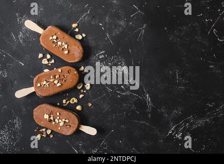 Milchschokolade Eis am Stiel mit Haselnüssen. Eis am Stiel bedeckt mit Schokolade, Stöcken und schwarzem Steinmarmorhintergrund. Platz für Text. Draufsicht. Stockfoto