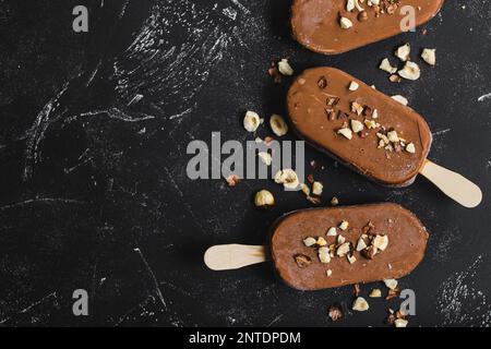 Milchschokolade Eis am Stiel mit Haselnüssen. Nahaufnahme. Eis am Stiel bedeckt mit Schokolade, Stöcken und schwarzem Steinmarmorhintergrund. Platz für Text. Stockfoto