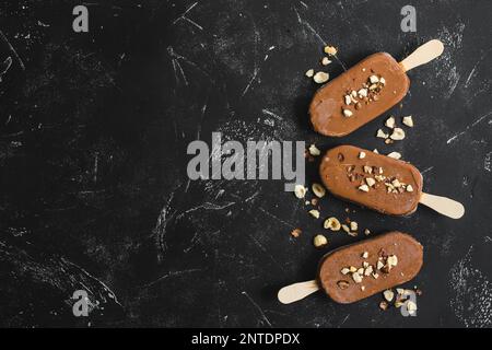 Milchschokolade Eis am Stiel mit Haselnüssen. Eis am Stiel bedeckt mit Schokolade, Stöcken und schwarzem Steinmarmorhintergrund. Platz für Text. Draufsicht. Stockfoto