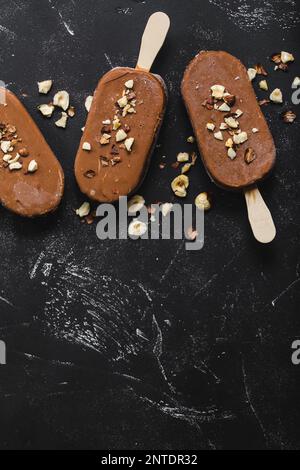 Milchschokolade Eis am Stiel mit Haselnüssen. Nahaufnahme. Eis am Stiel bedeckt mit Schokolade, Stöcken und schwarzem Steinmarmorhintergrund. Platz für Text. Stockfoto