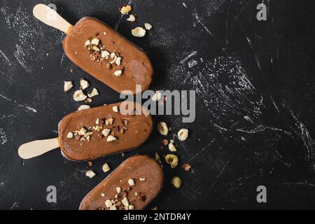 Milchschokolade Eis am Stiel mit Haselnüssen. Nahaufnahme. Eis am Stiel bedeckt mit Schokolade, Stöcken und schwarzem Steinmarmorhintergrund. Platz für Text. Stockfoto