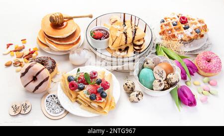 Süßer Ostertisch. Pfannkuchen, Crepes, Waffeln und Donuts mit frischen Beeren, Nüssen und Belag. Ostern traditionelle, natürliche bunte Eier. Stockfoto