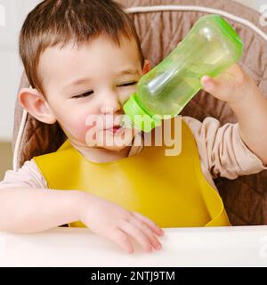 Glückliches Kleinkind, das aus einer Flasche trinkt, während es auf einem Hochstuhl sitzt. Der Junge im Lätzchen isst in der Küche zu Mittag. Ein Kind im Alter von einem Jahr sechs Stockfoto