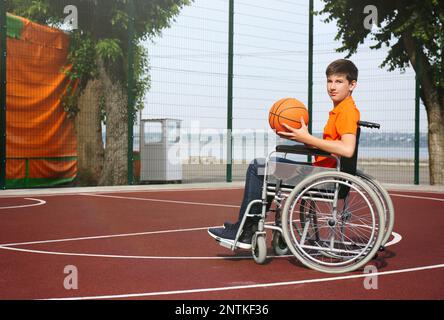 Behinderter Teenager im Rollstuhl mit Basketballball im Freien Stockfoto