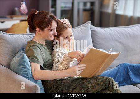 Seitliches Porträt einer Militärfrau mit Tochter zu Hause, die zusammen ein Buch liest und sich auf dem Sofa entspannt Stockfoto