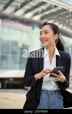 Porträt, attraktive und selbstbewusste asiatische Millennials-Geschäftsfrau in legerem Anzug, die ihr Smartphone außerhalb des Firmengebäudes verwendet. Stockfoto