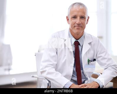 Ich bin hier, um zu helfen. Porträt eines männlichen Arztes, der in seinem Büro sitzt. Stockfoto
