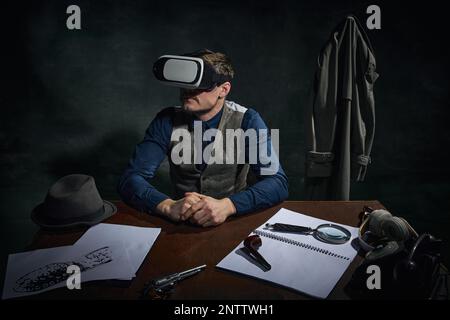 Professioneller Detektiv in klassischer Kleidung sitzt am Tisch in VR-Brille vor dunkelgrünem Vintage-Hintergrund. Untersuchung. Moderne Technologien Stockfoto