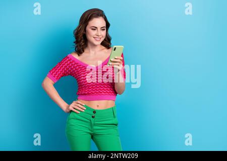 Foto von positivem braunem Haar lächelndes fröhliches Mädchen trägt ein pinkfarbenes Oberteil und durchsucht weitere youtube-Videos auf dem Smartphone isoliert auf blauem Hintergrund Stockfoto