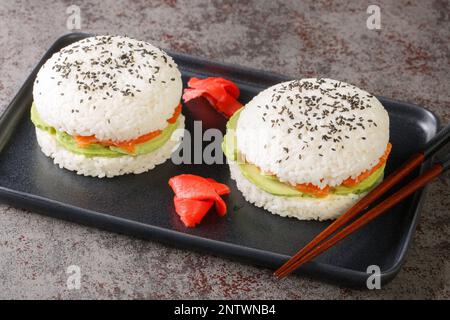 Reis-Sushi-Burger mit geräuchertem Lachs, Avocado und schwarzem Sesam, serviert auf einem Teller mit Holzstäbchen aus nächster Nähe. Horizontal Stockfoto