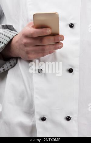 Ein professioneller Koch oder Koch hält ein Smartphone in der Hand. Im Hintergrund ist seine offizielle Uniform, weiß mit zwei parallelen schwarzen Knöpfen, ein SIG Stockfoto