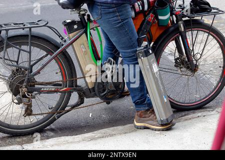 Lithium-Ionen-E-Bike-Batterien, die von einem Lieferboten getragen werden; eine auf dem E-Bike und eine Ersatzbatterie an seinem Fuß. Stockfoto