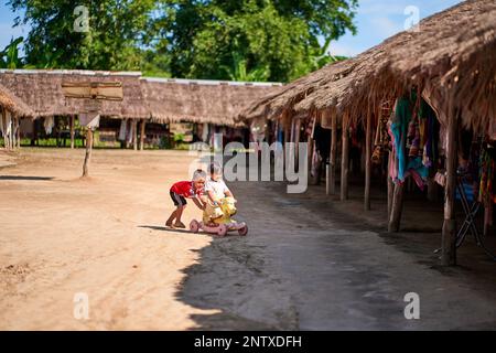 Zwei kleine Jungen spielen mit einem bunten Kinderfahrrad in einer ländlichen Umgebung in Thailand. Eine fröhliche Atmosphäre, die die Freude des Kindheitsspiels einfängt Stockfoto