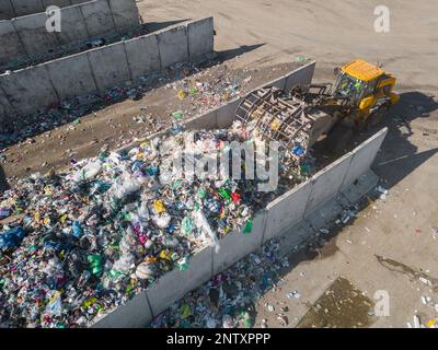 Gelber Radlader mit angehobenem Schrottgreifer, der sich im Bereich des Recyclingzentrums bewegt, während er entsorgten Abfall verarbeitet Stockfoto