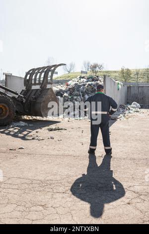 Ein Mitarbeiter des Recyclingzentrums in dunkelblauer Arbeitskleidung, der sich einen unsortierten Müllhaufen ansieht, ist gerade in der Materialrückgewinnungsanlage eingetroffen Stockfoto