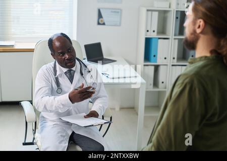 Selbstbewusster reifer Arzt mit medizinischem Dokument, das mit dem jungen Patienten spricht, der während der Sprechstunde in Kliniken vor ihm sitzt Stockfoto