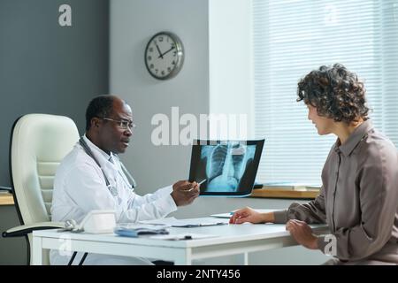Reifer Arzt, der auf das Röntgenergebnis der Lunge zeigt, während er es der reifen Patientin erklärt und die Diagnose während der ärztlichen Konsultation stellt Stockfoto