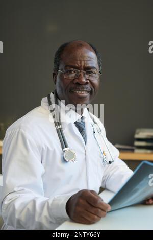 Erfahrener afroamerikanischer Radiologe mit Röntgenbildern der Lunge, wenn der Patient lächelnd vor die Kamera schaut, während er am Arbeitsplatz sitzt Stockfoto