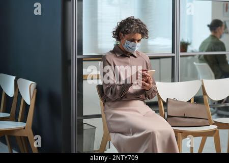Reife Patientin in Freizeitkleidung und Schutzmaske, die SMS auf dem Smartphone schreibt, während sie auf dem Stuhl sitzt und darauf wartet, dass sie zum Arzt geht Stockfoto