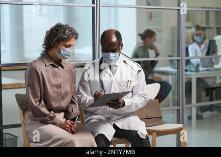 Reifer Arzt und Patientin in Schutzmasken, die im Wartezimmer der Klinik Rezepte diskutieren Stockfoto