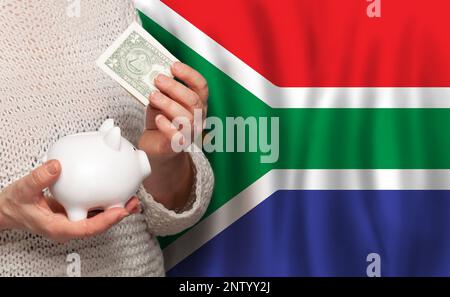 Südafrikanische Frau mit Geldbank auf dem Hintergrund der südafrikanischen Flagge. Punkte, Pensionsfonds, Armut, Wohlstand, Altersvorsorge Stockfoto