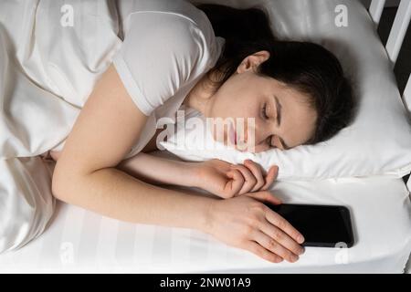 Eine 30-jährige Frau schläft mit ihrem Handy neben einem Kissen. Telefonsüchtig, immer online Stockfoto