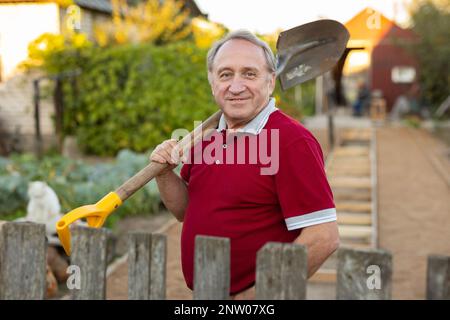 Ein erwachsener Mann stand entspannt neben dem Zaun, mit einer Schaufel im Garten, die lächelte Stockfoto