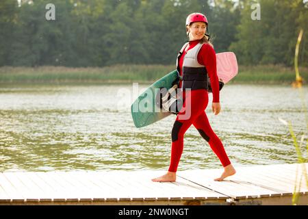 Frau in Neoprenanzug, Helm und Schwimmweste, die mit einem Wakeboard auf einem Pier läuft. Sonniger Sommertag. Sicherheit im Sport. Wassersport in Finnland. Versicherungskonz Stockfoto