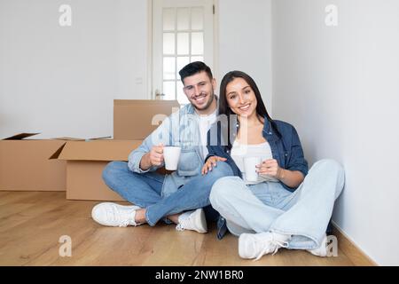 Glückliches junges Paar, das nach Hause zieht, zwischen Pappkartons sitzt und Kaffee trinkt Stockfoto