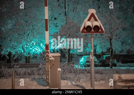 Semaphor oder Signal für entgegenkommenden Zug mit Barrieren in der Nacht. Neigungsüberquerung mit Lichtern und Barrieren bei Schneewetter in der Nacht. Stockfoto