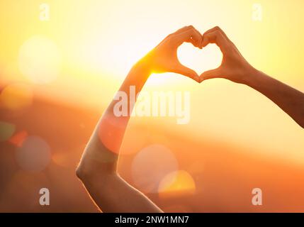 Verliebt in die Landschaft. Aufnahme von Händen einer nicht identifizierbaren Frau, die über einer Sonnenuntergangslandschaft eine Herzform bilden. Stockfoto