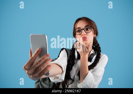 Ein lächelndes europäisches Mädchen mit Zöpfen und einem Rucksack in Brille und Kopfhörern macht Fotos am Telefon Stockfoto