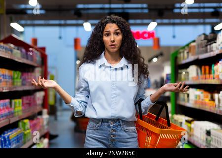Eine besorgte junge lateinamerikanische Frau steht in einem Supermarkt zwischen den Regalen mit Waren, hält einen Korb, spreizt ihre Hände, schaut in die Kamera. Produkt nicht gefunden, nicht auf Lager. Stockfoto