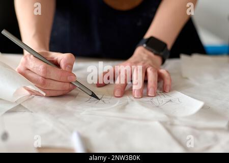 Die Hände einer kreativen Schneiderin zeichnen mit Bleistift Skizzen neuer Kleidung auf weißem Papiermuster und bereiten sie für die weitere Arbeit vor Stockfoto