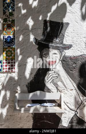 Straßenkunst eines Mannes mit einem Hut, der ein Glas Rotwein in einer friedlichen Straße von Grottaglie, Apulien, Italien hält Stockfoto