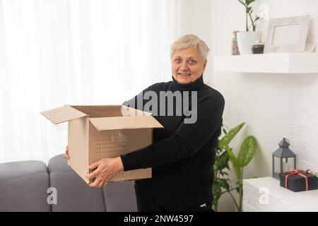 Lächelnde ältere Frau erhält zu Hause ein Postpäckchen, glückliche alte Großmutter hält den Karton offen und sitzt auf dem Sofa im Wohnzimmer Stockfoto