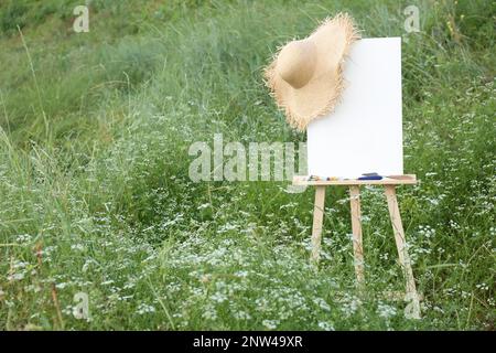 Hölzerne Staffelei mit leerer Leinwand, Malausrüstung und Hut auf der Wiese. Platz für Text Stockfoto