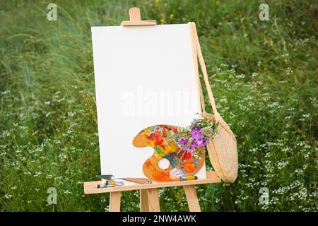 Hölzerne Staffelei mit leerer Leinwand, Malmaschinen und Blumen auf der Wiese. Platz für Text Stockfoto