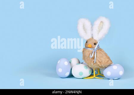 Osterküken mit Hasenohren und pastellfarbenen Ostereiern auf blauem Hintergrund mit Kopierbereich Stockfoto