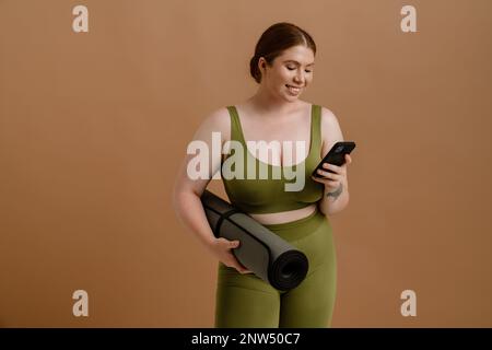 Ein junges, wunderschönes, lächelndes Mädchen in Übergröße im Trainingsanzug mit Telefon und Yogamatte, das auf einem isolierten braunen Hintergrund steht Stockfoto