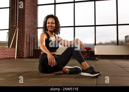 Attraktive afroamerikanische Frau in Sportbekleidung, die in die Kamera schaut und lächelt, während sie in einem Crossfit-Fitnessstudio auf dem Boden sitzt Stockfoto