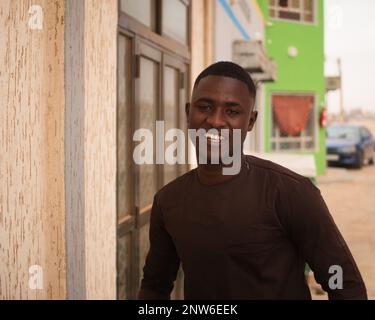 Ein junger afrikanischer Mann in brauner Kleidung lächelt vor der Kamera in den Straßen von Nouakchott, Mauretanien Stockfoto