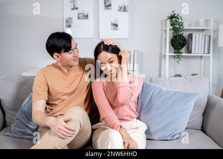 Ein junges asiatisches, glückliches Paar, das lächelt und auf der Couch im Wohnzimmer zu Hause sitzt. Freund und Freundin umarmen und entspannen Stockfoto