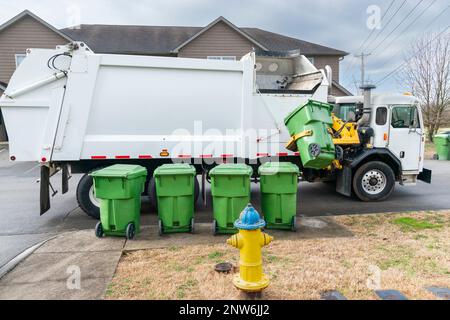Horizontale Aufnahme eines automatisierten Müllwagens, der den Müllcontainer auf dem Bordstein aufnimmt. Vier weitere Mülltonnen stehen in Schlange und warten darauf, dass sie dran sind. Stockfoto